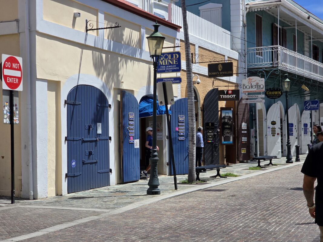 Street scene in St. Thomas showing several side-by-side jewelry stores with arched doors, hanging signs, and a few shoppers walking past on a bright day.