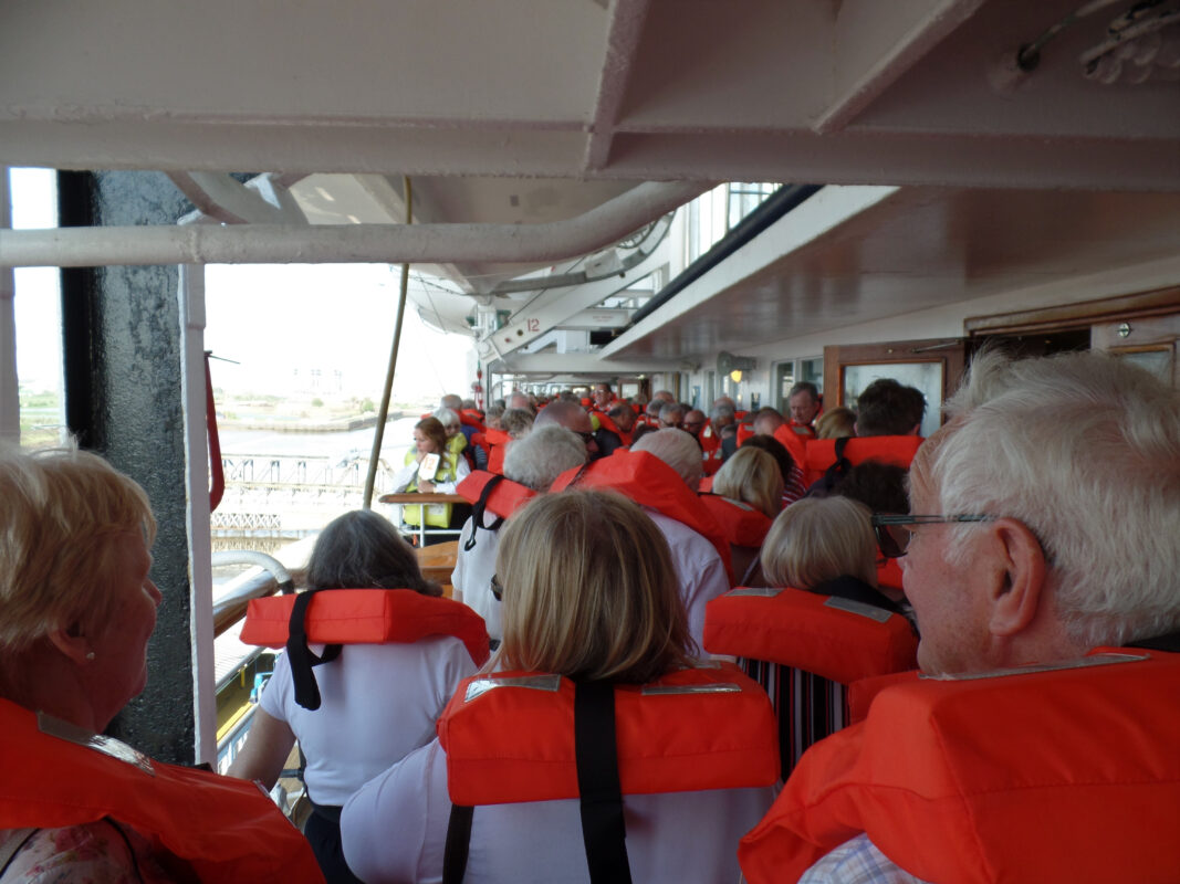 Passengers wearing bright orange life jackets stand in a crowded line along an outdoor ship deck corridor during a safety drill.