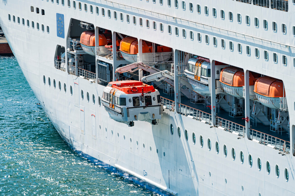 Close-up of a white cruise ship’s side showing multiple orange lifeboats stored in davits and one enclosed lifeboat suspended above the water.