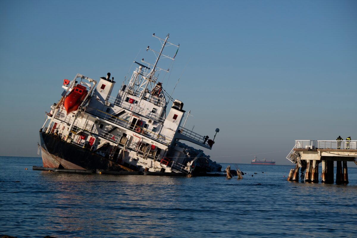 A damaged vessel leans heavily in the water beside a dock, with debris floating nearby and a distant ship on the horizon.