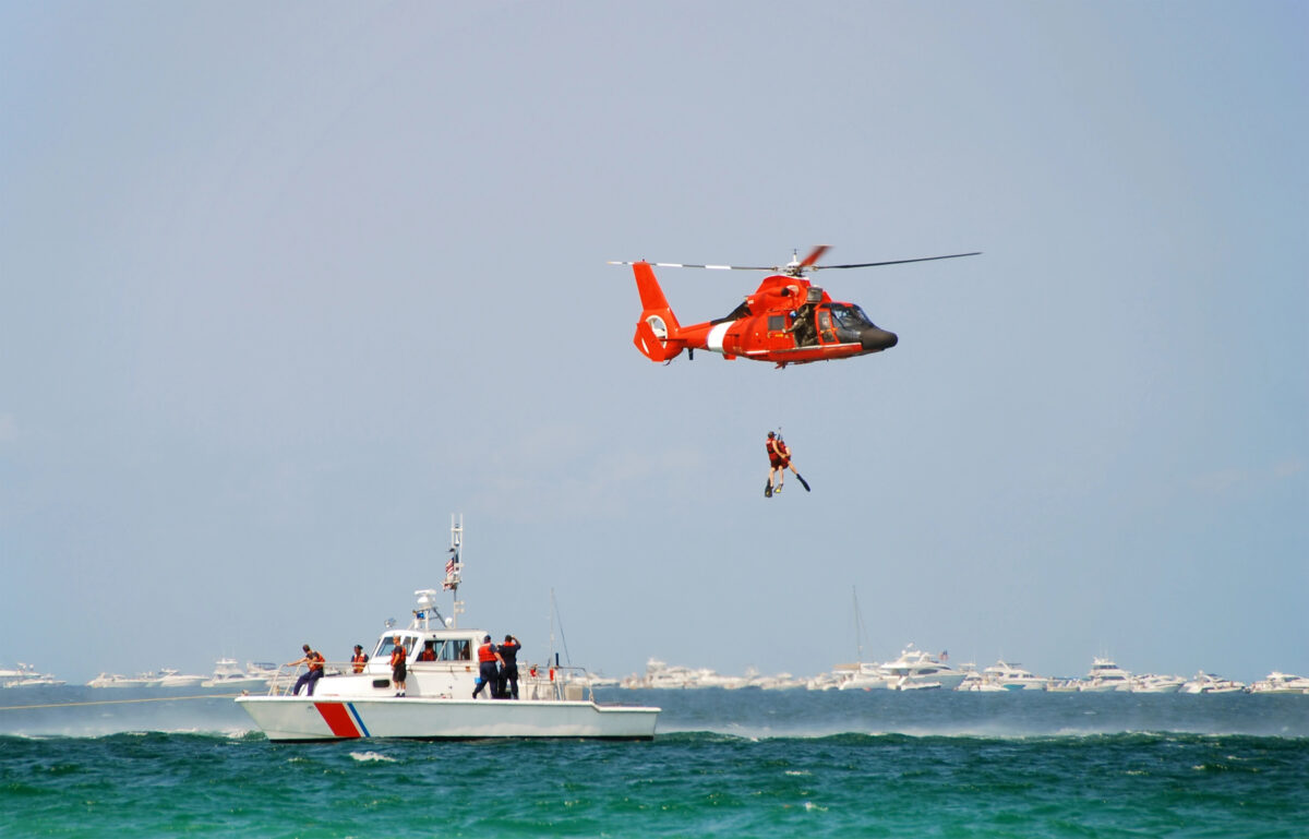 Coast Guard style helicopter rescue above open water with a swimmer being lifted and a rescue vessel nearby.