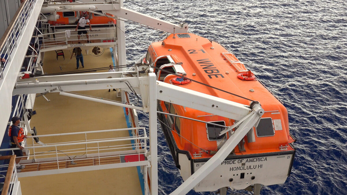 Crew members on a cruise ship deck next to an orange lifeboat being lowered over the water during an emergency equipment drill.