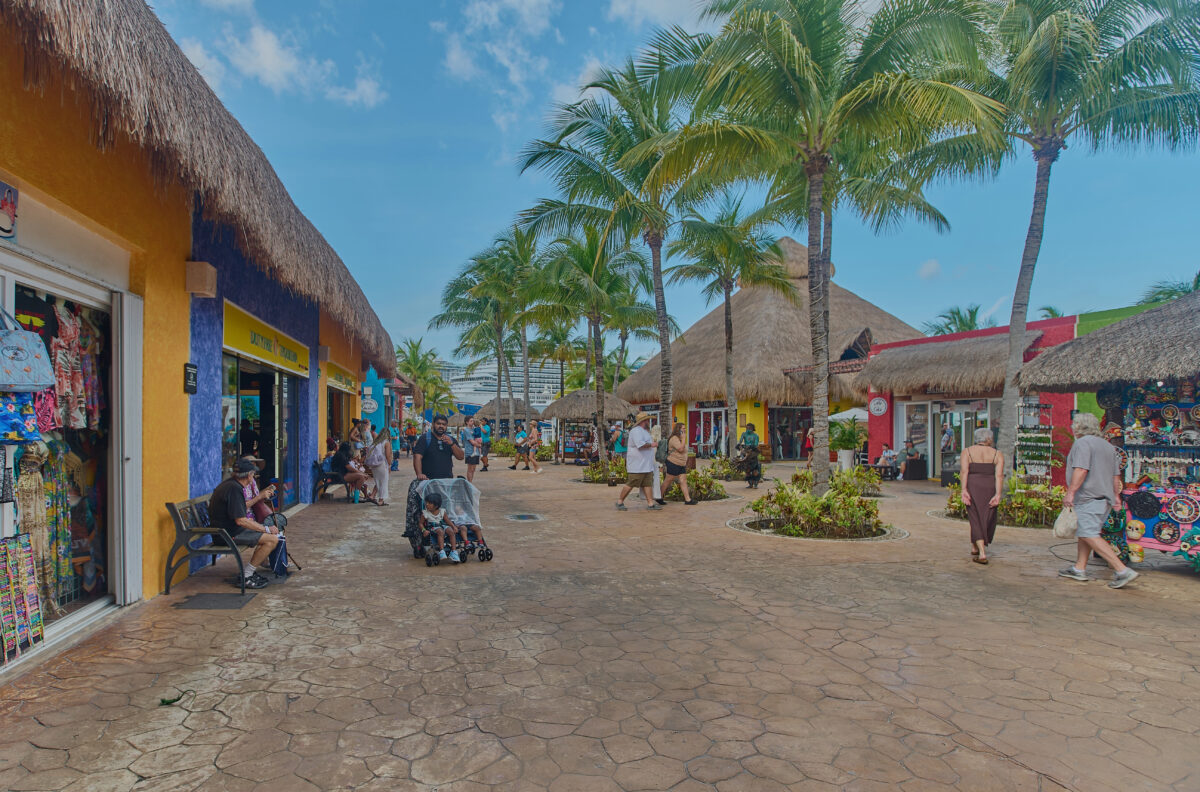 Cozumel pier shopping area showing a row of bright shops on the left, souvenir stands on the right, and families strolling under tall palm trees with a cruise ship visible in the background.
