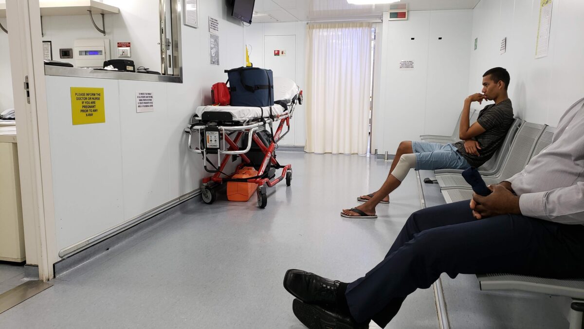 Interior of an onboard medical clinic with bench seating, a patient waiting, and emergency equipment on a gurney.