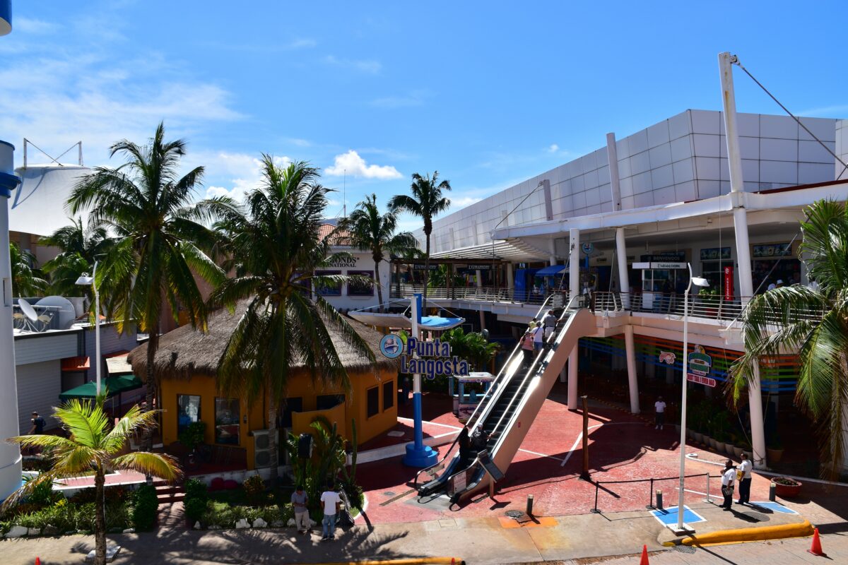 Cozumel cruise port at Punta Langosta, showing the shopping complex and elevated walkway, with palm-lined plaza, escalator, and a few people near the entrance.