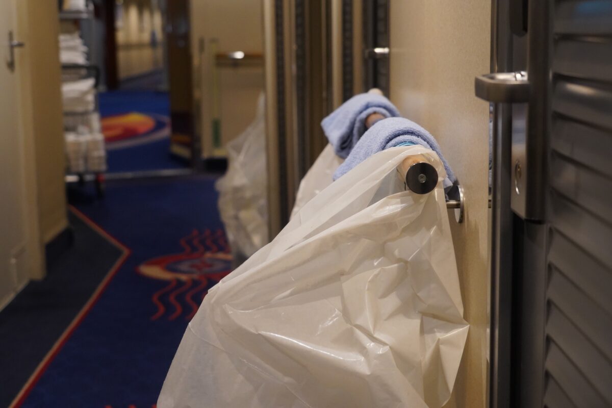 Cruise ship corridor with a housekeeping trolley, folded towels, and a white laundry bag outside stateroom doors.