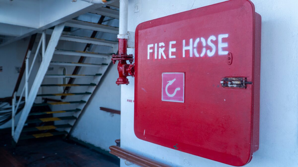 Red fire hose cabinet mounted on a cruise ship wall near a metal stairwell.