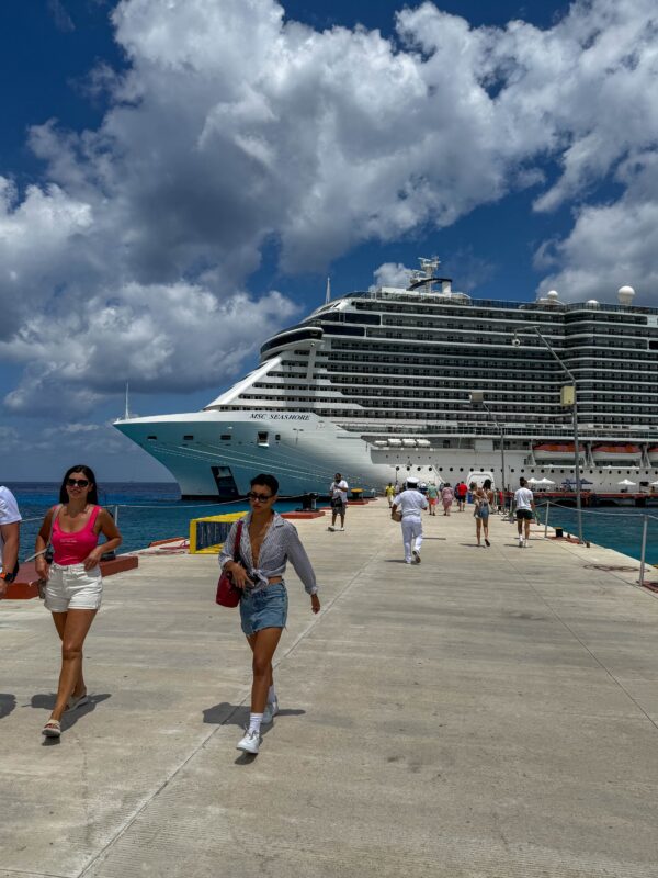People leaving a docked cruise ship and heading down the pier on a sunny port day, with the ship dominating the background.