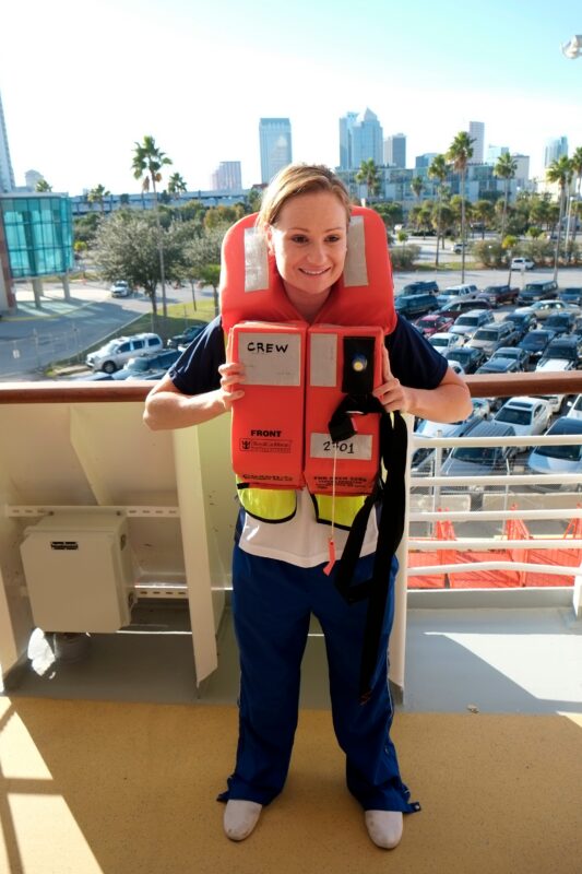 Woman wearing an orange life jacket on a cruise ship deck near a railing, illustrating muster drill safety instructions.