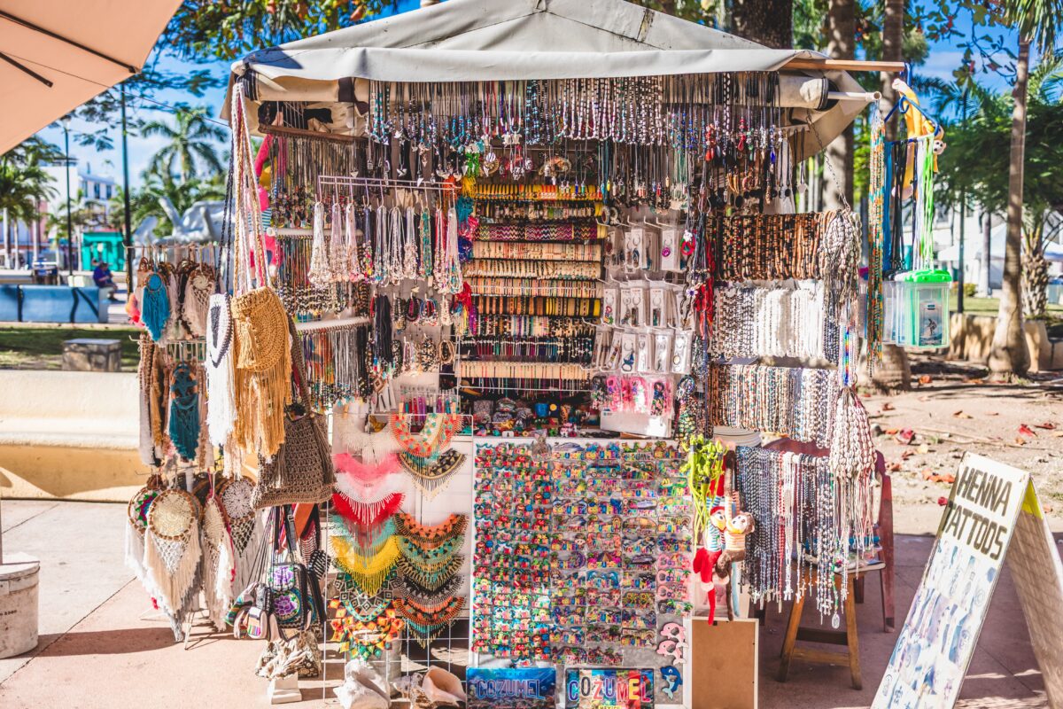 Outdoor souvenir stall in Cozumel covered with hanging necklaces, bracelets, earrings, woven bags, and colorful trinkets under a canopy on a sunny day.