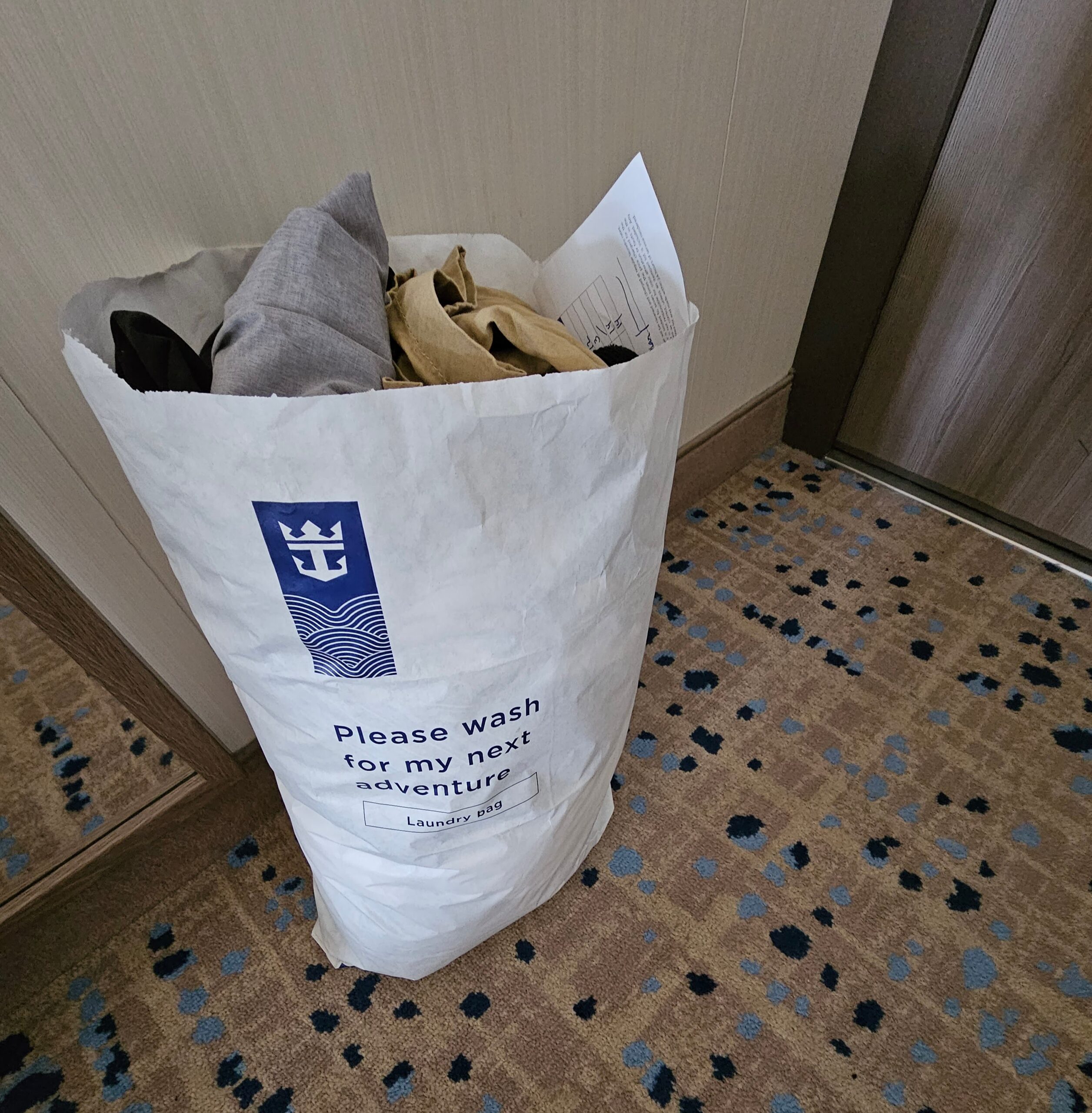 Large cruise laundry bag holding used clothing in a stateroom with patterned carpet and wood trim.