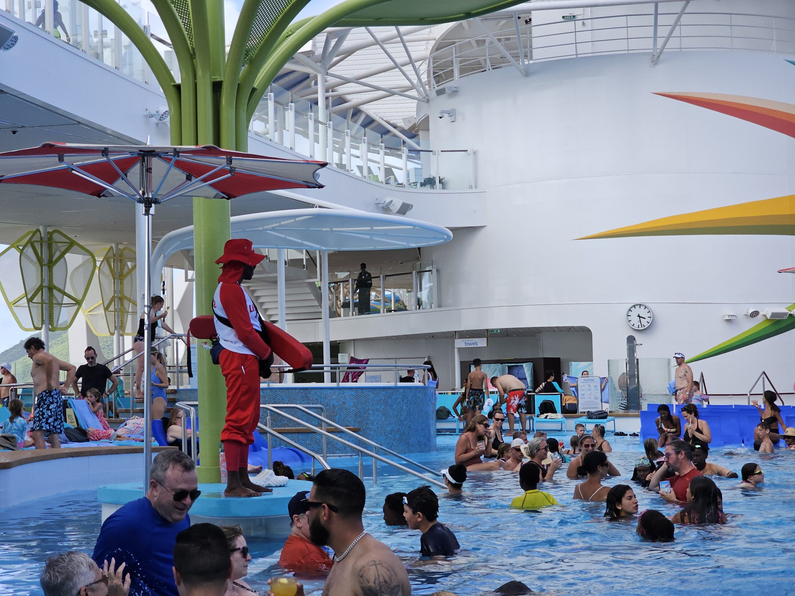 Lifeguard in red uniform standing on a raised platform beside a busy cruise ship pool filled with families.