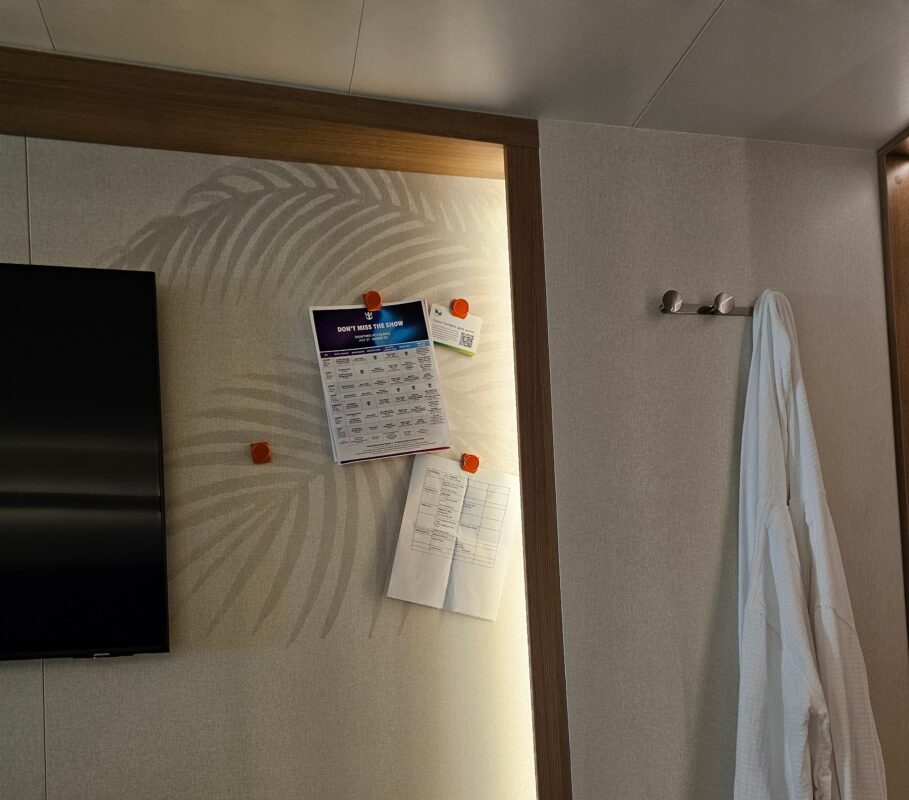 Cruise cabin wall with orange magnets holding daily schedules and papers beside a television and hanging robe.
