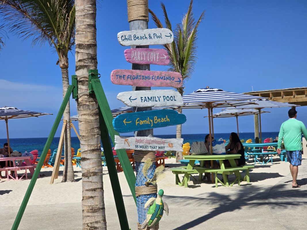 Colorful wooden sign at Royal Beach Club Paradise Island with arrows for Chill Beach, Party Cove, Family Beach, and other attractions near the sand.