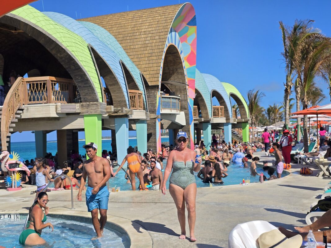 Crowded pool scene at Party Cove in Royal Beach Club Paradise Island, with guests in the water and colorful arched buildings behind them.