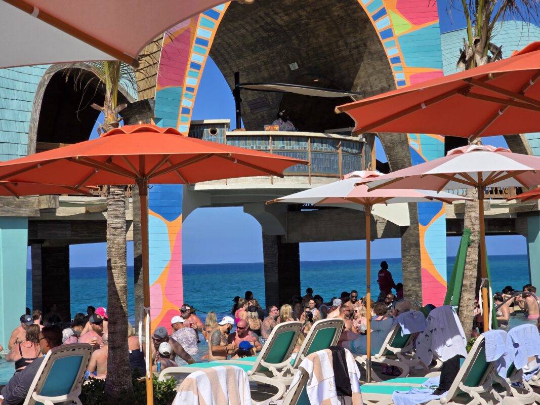 View of Party Cove showing the raised DJ booth, poolside loungers, umbrellas, and guests gathered below at Royal Beach Club Paradise Island.