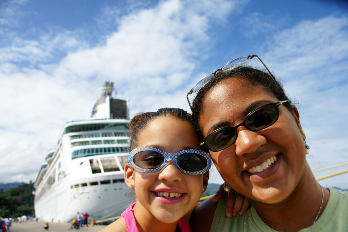 Mother and daughter posing for a selfie near a cruise ship docked at port under a blue sky.