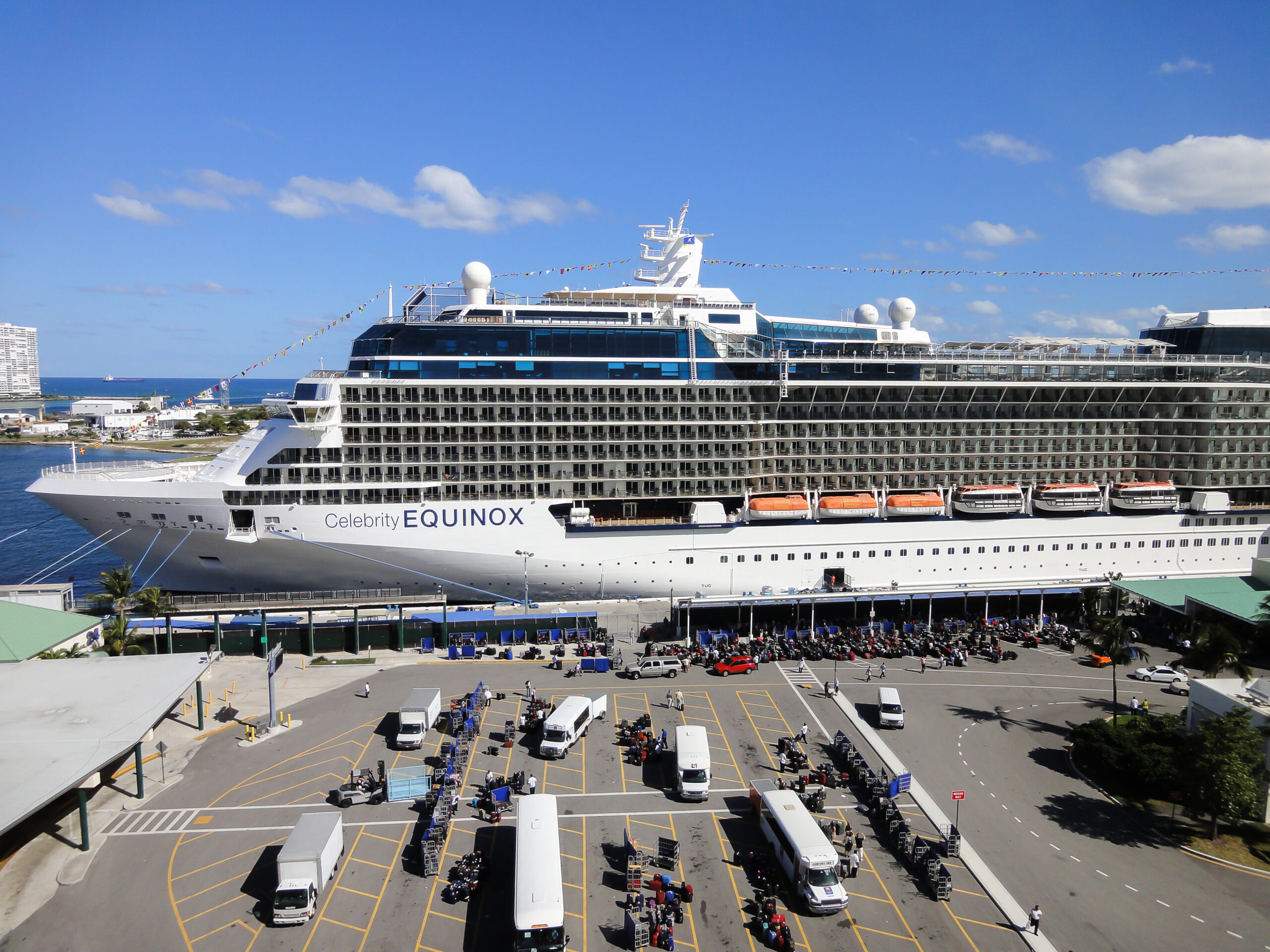 Wide view of Celebrity Equinox docked beside a cruise terminal with buses, carts, and embarkation activity on the ground below.