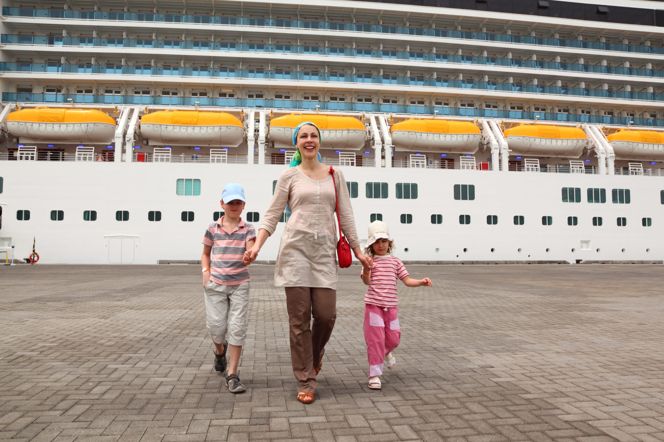Woman walking with two children in front of a large cruise ship while heading ashore in port.
