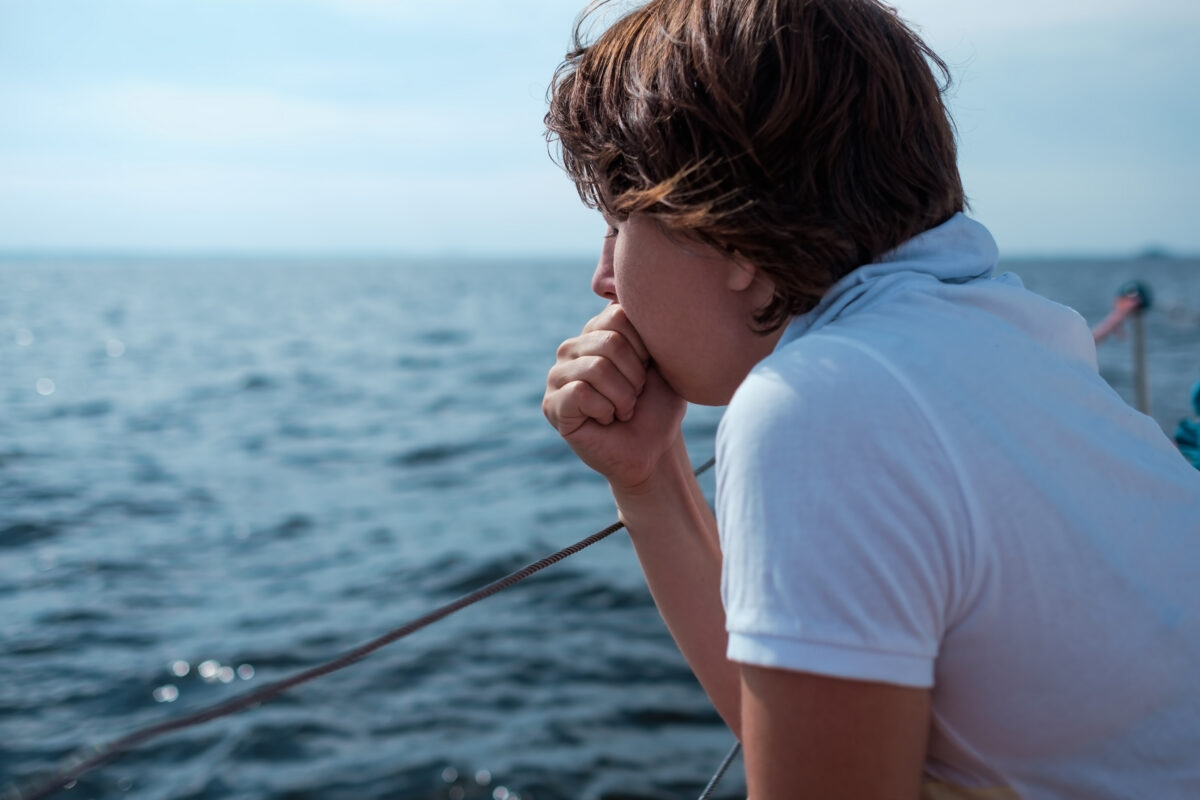 Young passenger gazing out at the water while standing along an outdoor deck railing with closed eyes and hand over mouth.