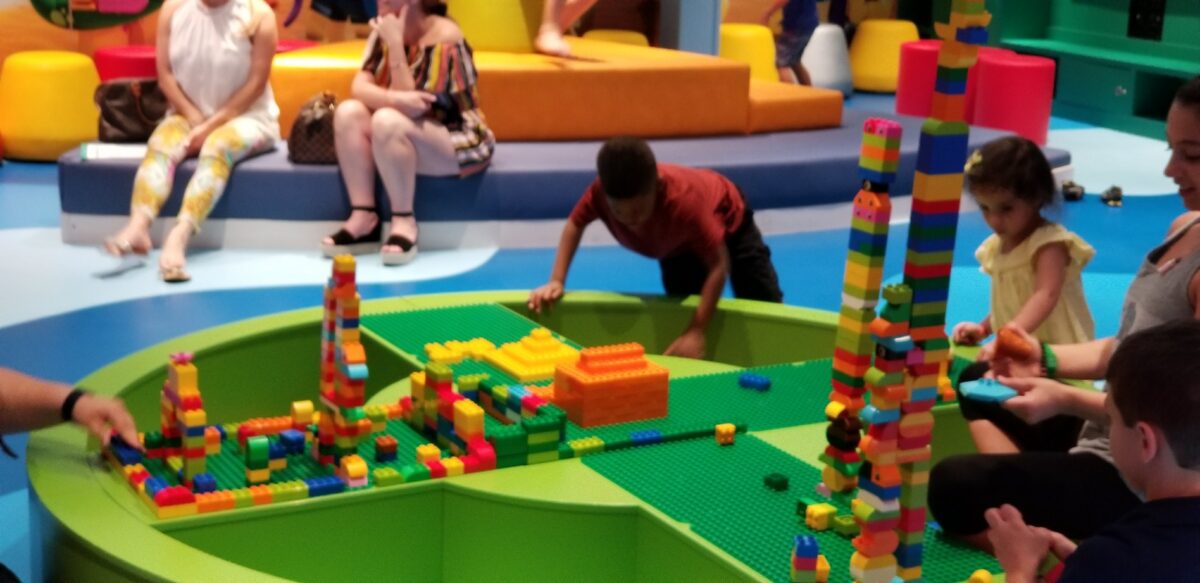 Children play with large colorful blocks in a Lego-themed cruise ship playroom while adults sit nearby.