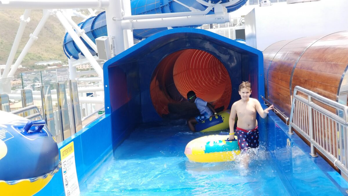 Boy steps out of a cruise ship water slide splash area while another child rides a tube inside the slide.