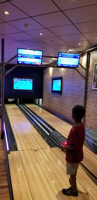 Young boy stands at a cruise ship bowling lane preparing to bowl, with screens and pins visible ahead.