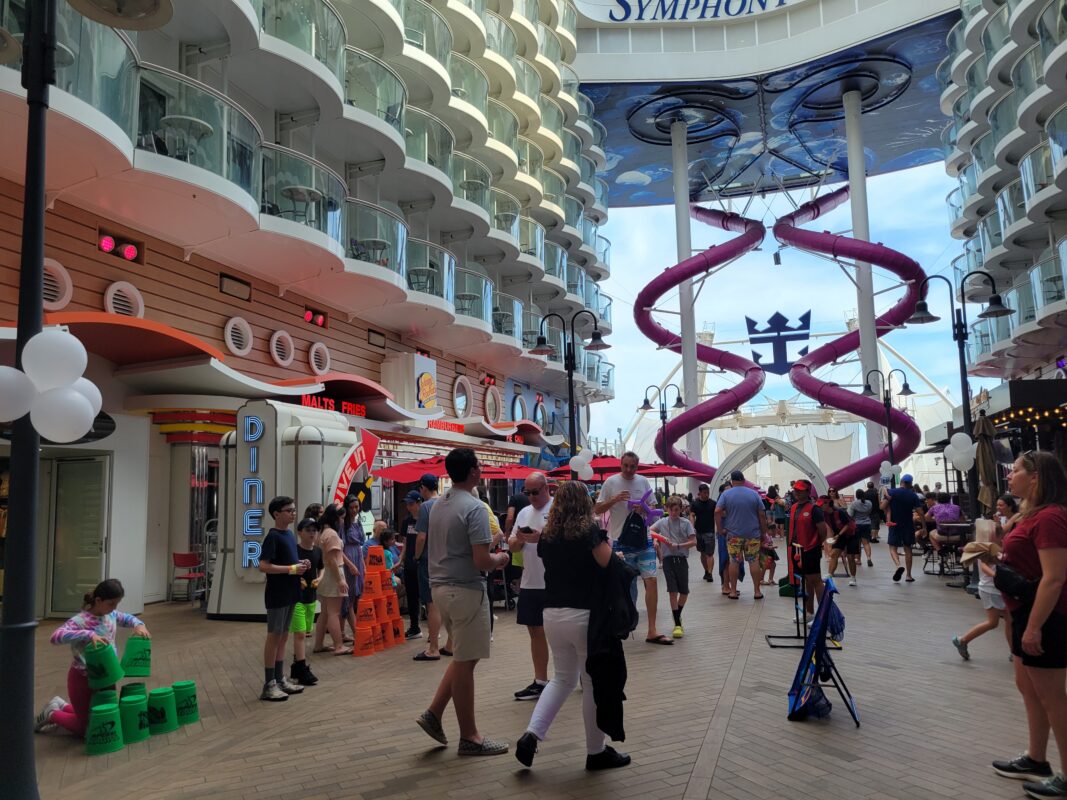 Families gather on a cruise ship boardwalk during an outdoor carnival, with games, shops, and a large purple slide.