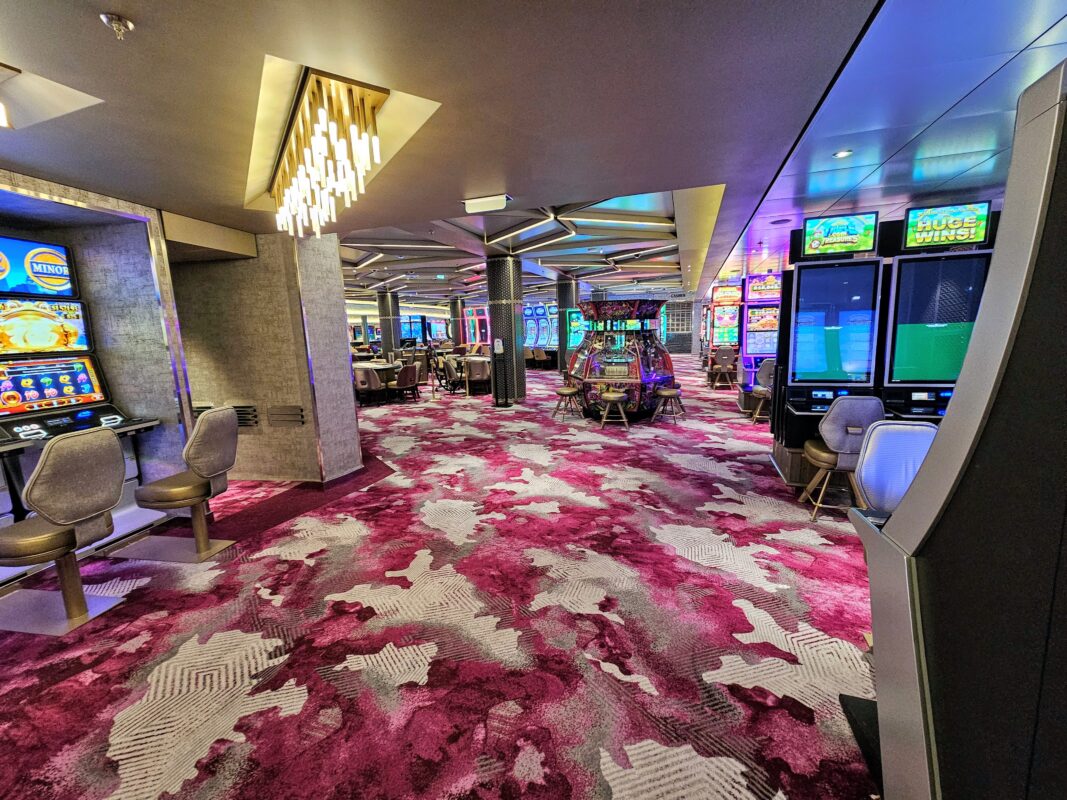 Cruise ship casino with slot machines, gaming tables, colorful patterned carpet, and bright ceiling lights.