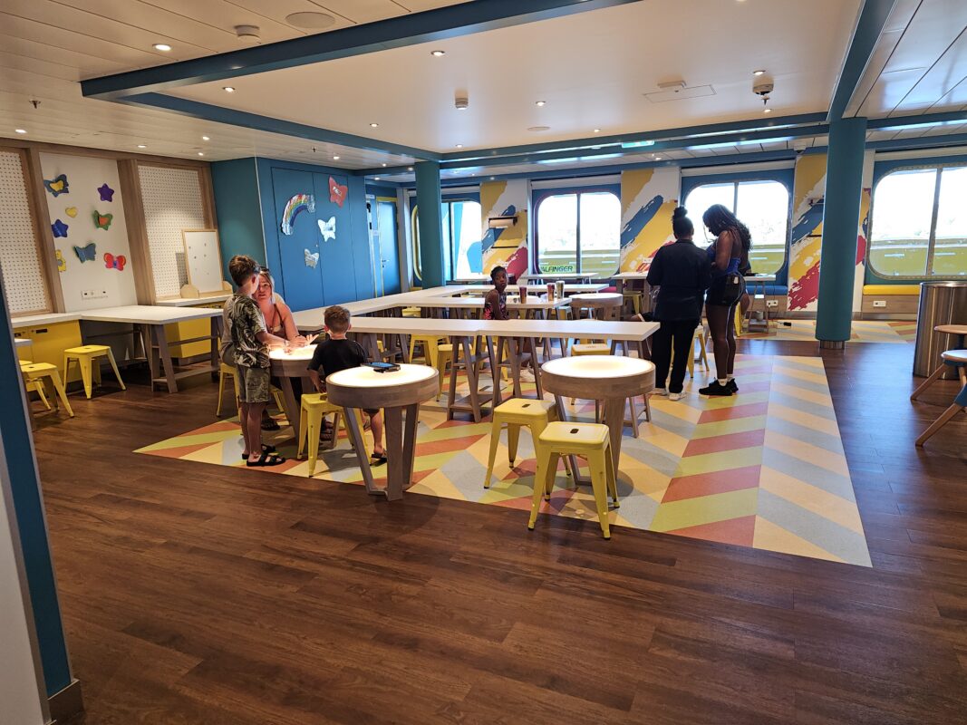 Kids and adults gather in a colorful cruise ship kids’ club room with tables, stools, and craft areas during open house.