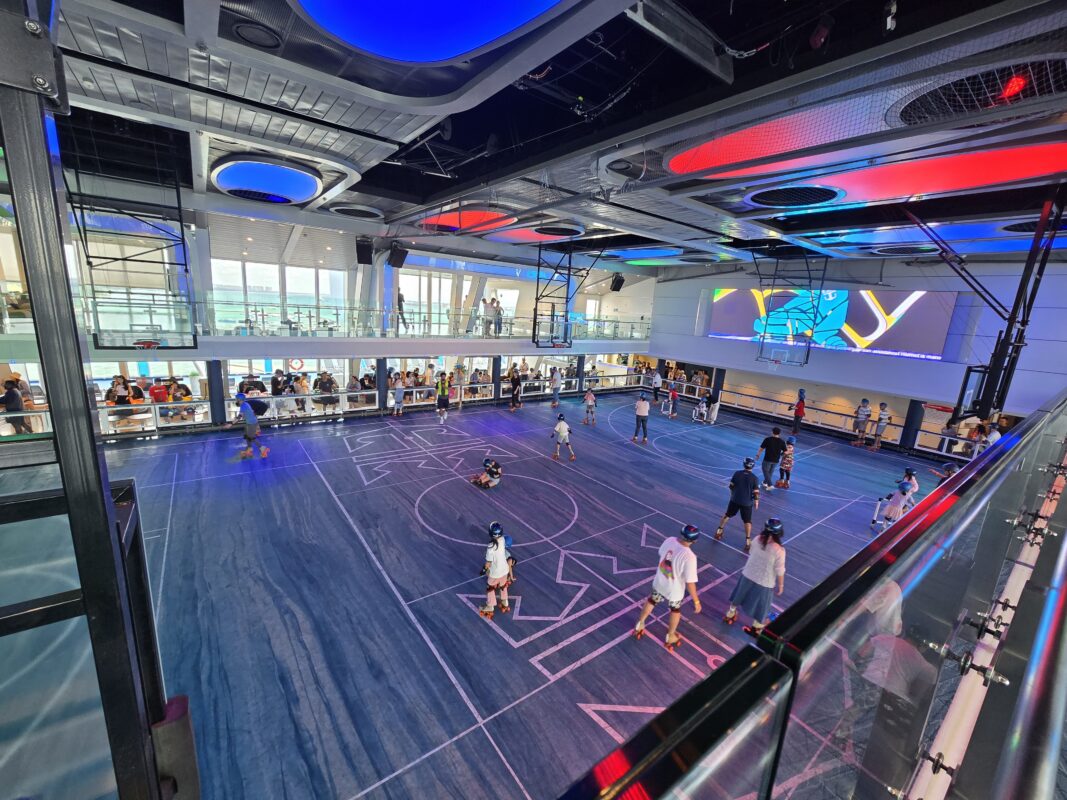 Passengers roller skating on an indoor sports court aboard a cruise ship, with colorful ceiling lights and ocean views through large windows.