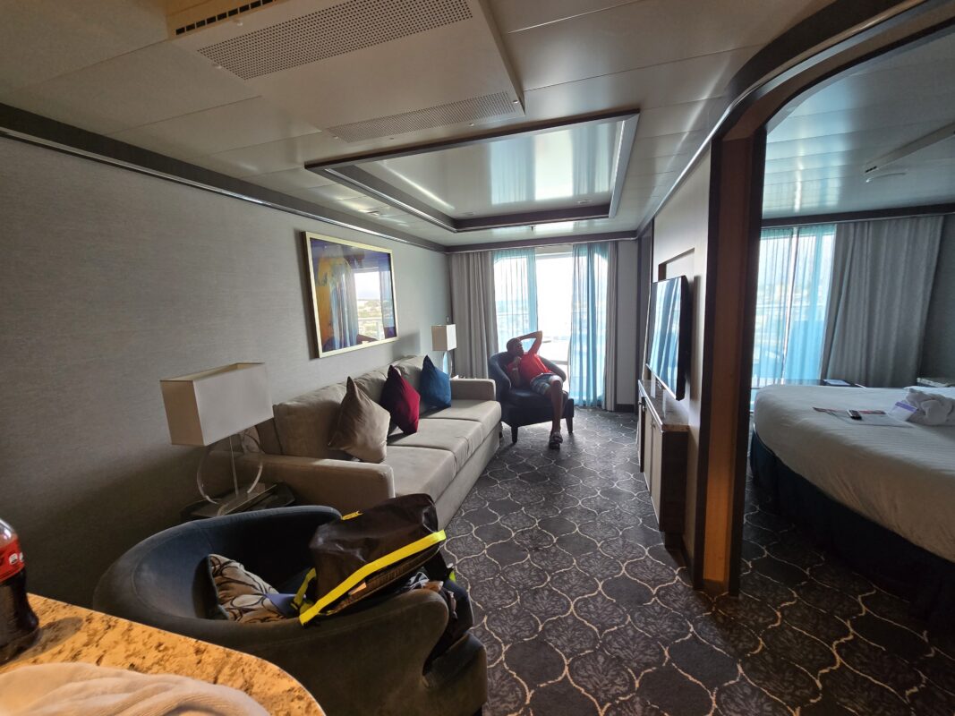 Roomy cruise suite featuring a couch, chairs, bed, TV, and balcony access, with a child seated near the window in the background.