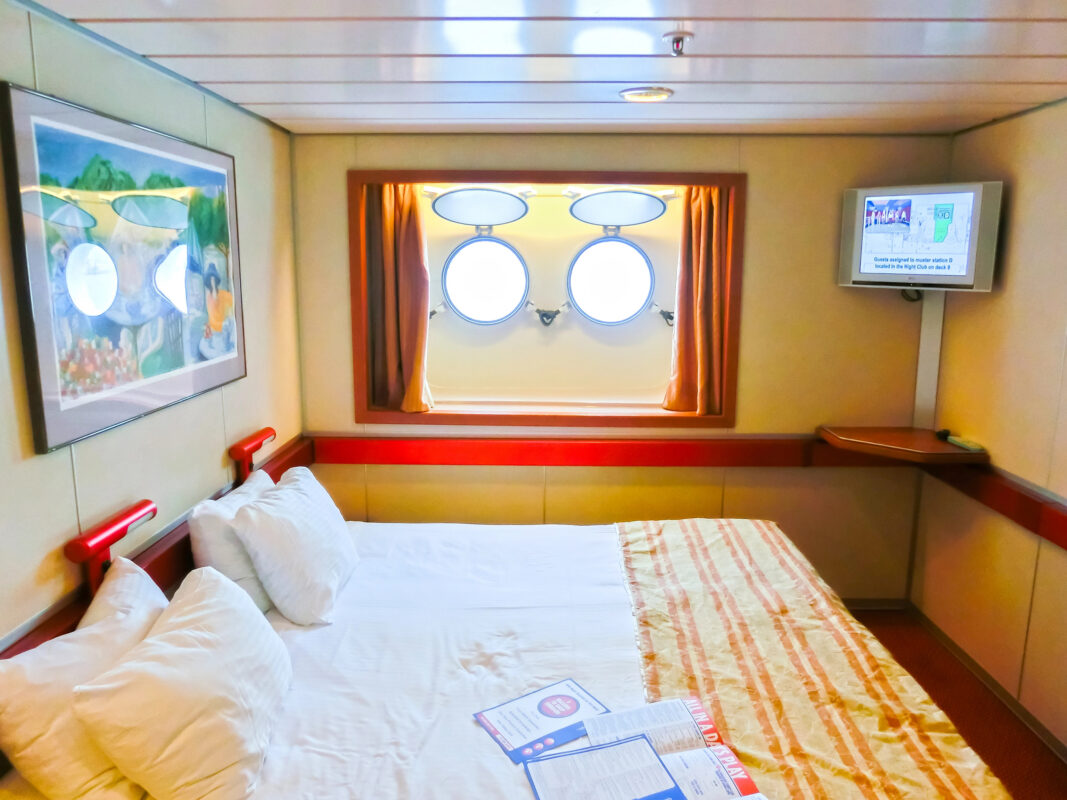 Basic cruise ship stateroom with a bed beneath two round porthole windows, warm-colored walls, a small TV, and limited floor space.