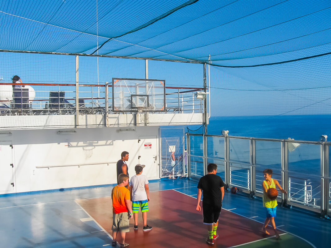 Group of passengers on a cruise ship basketball court enclosed by netting, with blue ocean visible beyond the railings.