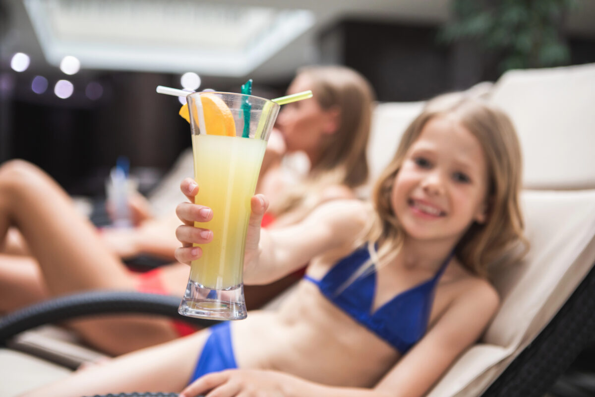 Young cruise passenger lounging poolside in a blue swimsuit and holding a yellow drink in a tall glass with fruit garnish.