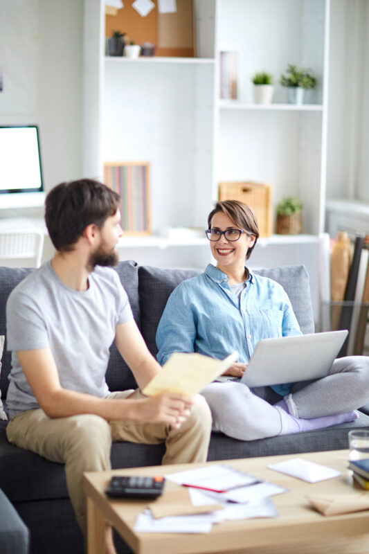 Two adults sitting on a couch with a laptop, papers, and a calculator nearby while discussing cruise expenses at home.