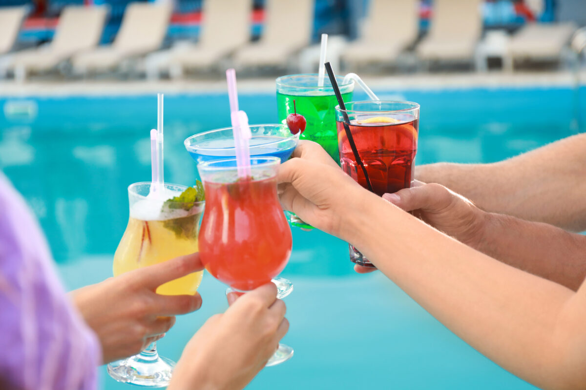 Several people holding colorful drinks near a cruise ship pool, with lounge chairs blurred in the background.