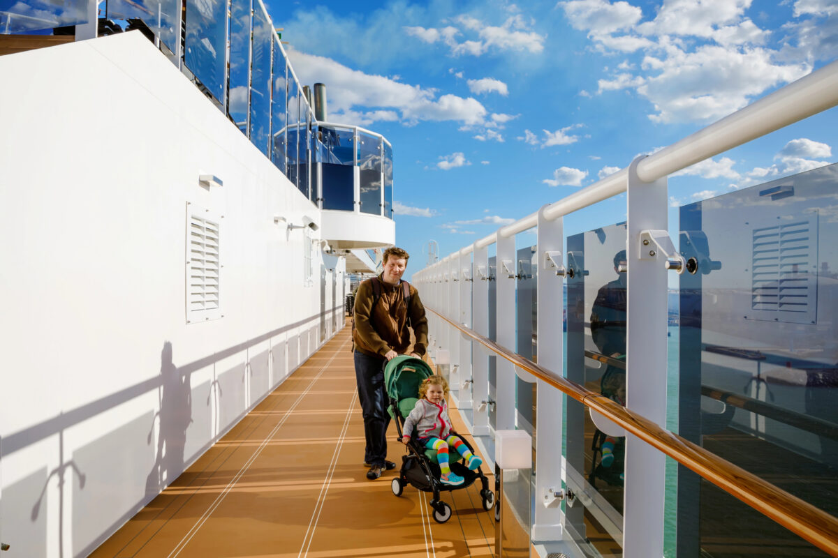 Adult pushes a toddler in a stroller along an outdoor cruise ship deck walkway.