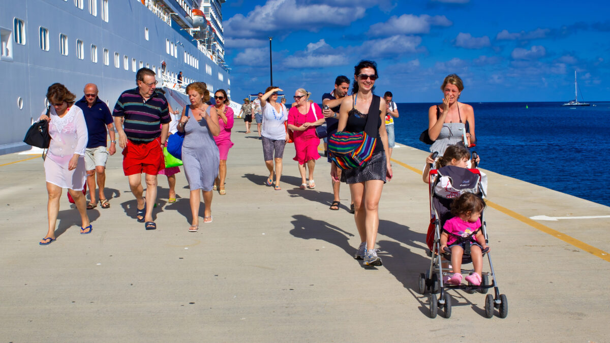 Families and other cruise passengers walking on a wide pier beside a cruise ship, including a parent pushing a stroller under a bright blue sky.