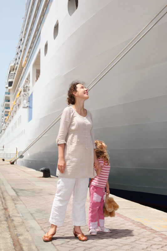 Woman and young girl holding hands beside a large docked cruise ship, with the child holding a teddy bear.