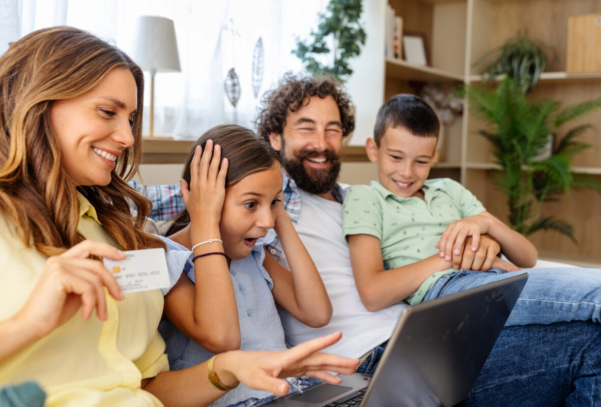 Parents and 2 children smile at a laptop during trip planning at home, with a credit card visible in the mother’s hand.