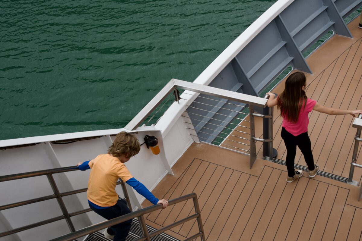 Two kids on a luxury family cruise descend a wide outdoor staircase on a cruise ship, with wood-look decking, metal railings, and water visible below.