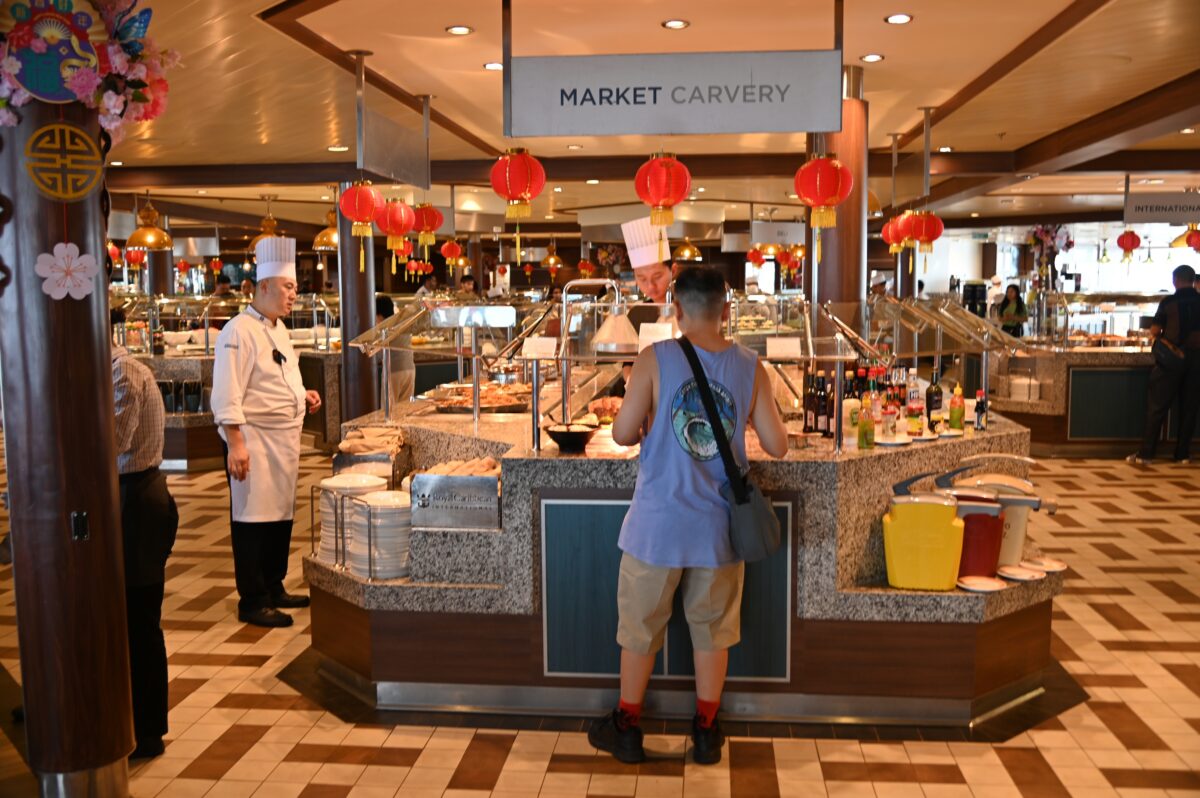 Passenger at a cruise ship buffet carving station with chefs behind the counter and red lantern decorations hanging overhead.