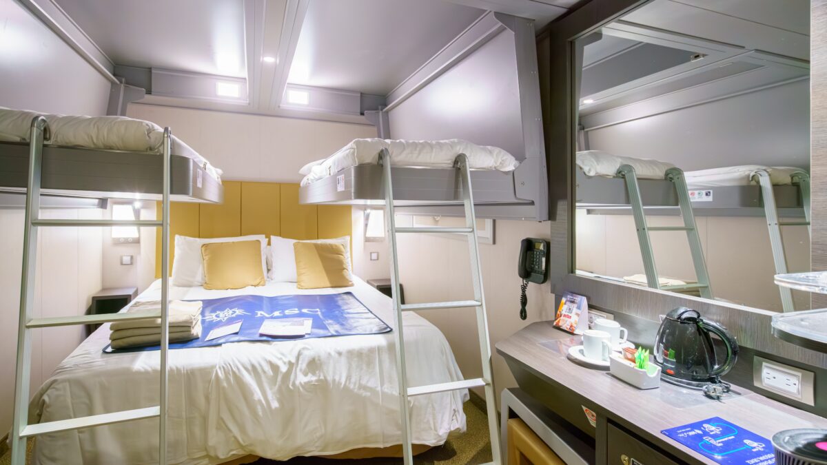 Modern cruise cabin with a bed beneath two overhead bunk beds, soft lighting, folded towels, and a desk area with a kettle and mugs.