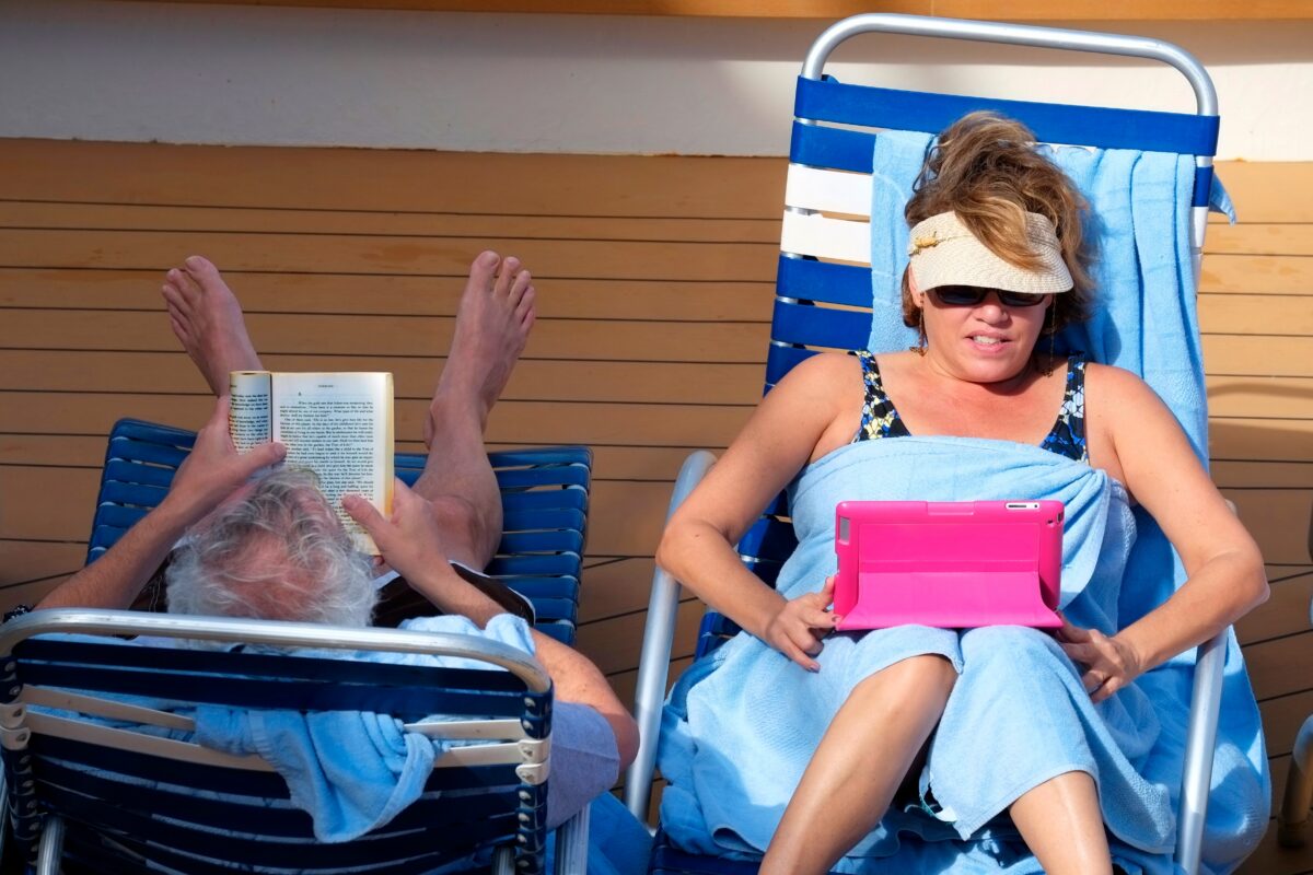 Woman using a tablet while reclining on a cruise deck chair next to another passenger reading a book in the sun.