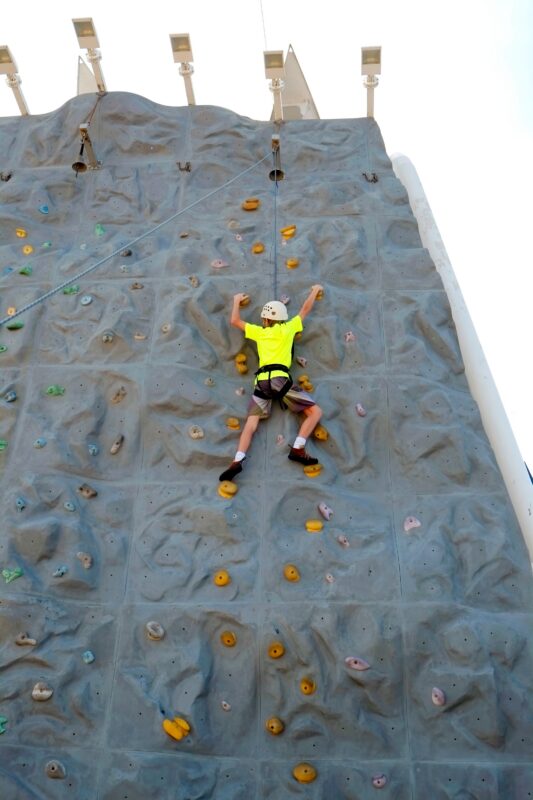 Young person scaling a cruise ship rock-climbing wall that is included in base fare cruise expenses and doesn't add to the fare.