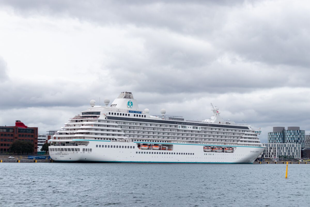 Crystal Serenity viewed from the side while docked near an urban waterfront, with gray water in the foreground and buildings in the background.