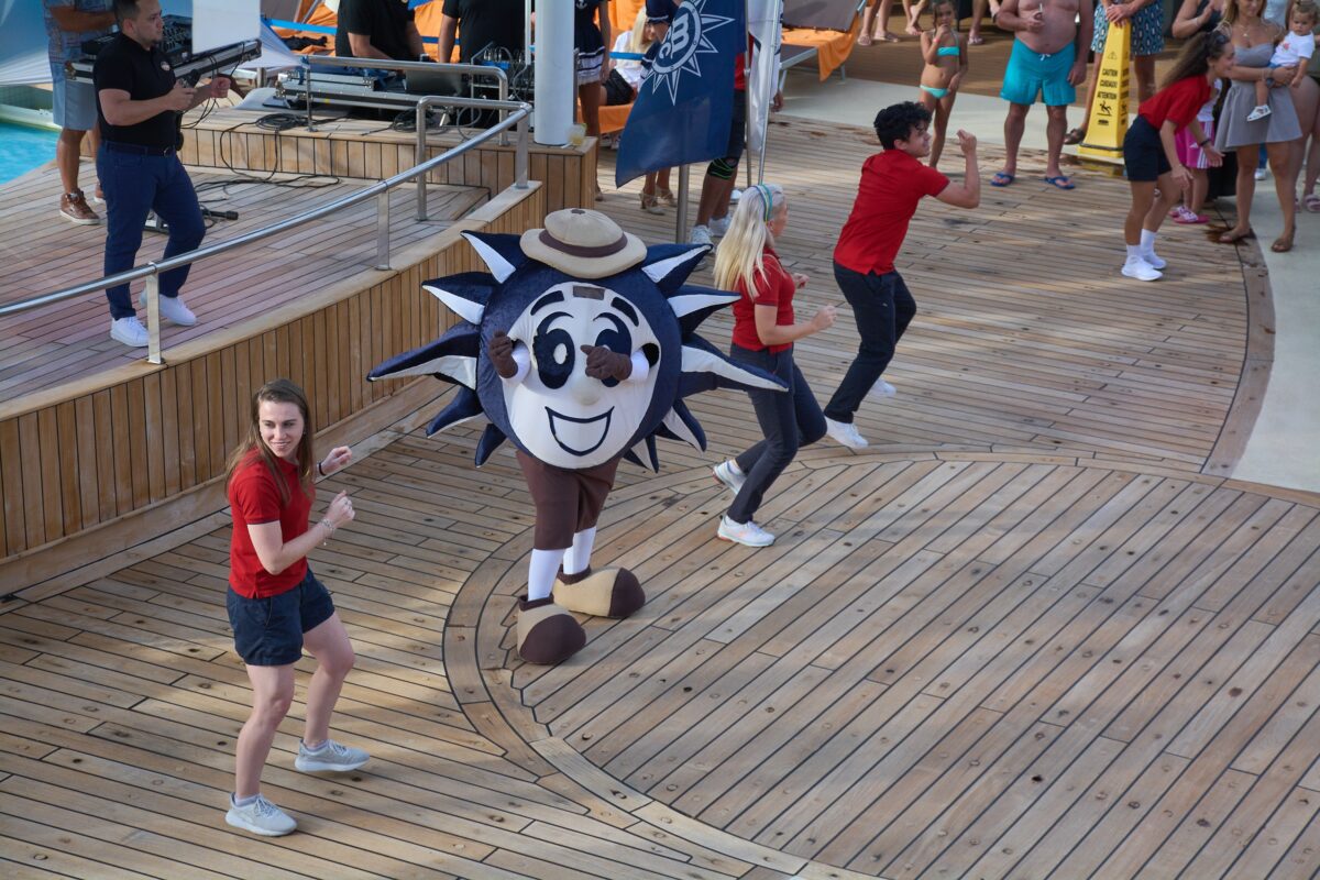 Cruise ship deck party with crew members dancing beside a smiling mascot character while passengers watch nearby.