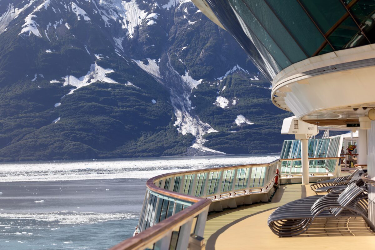 Alaska Family Cruise Deals | Empty outdoor deck on a cruise ship with rows of lounge chairs, glass wind screens, and a view of snowy mountains across the water.