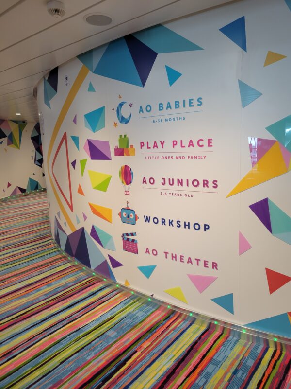 Colorful curved wall at the entrance to an exemplar cruise ship kids' club, displaying five labeled zones: AO Babies (6–36 months), Play Place (Little Ones and Family), AO Juniors (3–5 years old), Workshop, and AO Theater, with geometric triangle graphics and a multicolored striped carpet in the foreground.